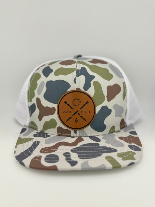 "Camo-patterned trucker hat with a white mesh back and a brown leather patch on the front. The patch features a circular design with two crossed arrows, a wreath, and the phrase 'Death to Life' engraved in the center, creating a bold and meaningful outdoor aesthetic."