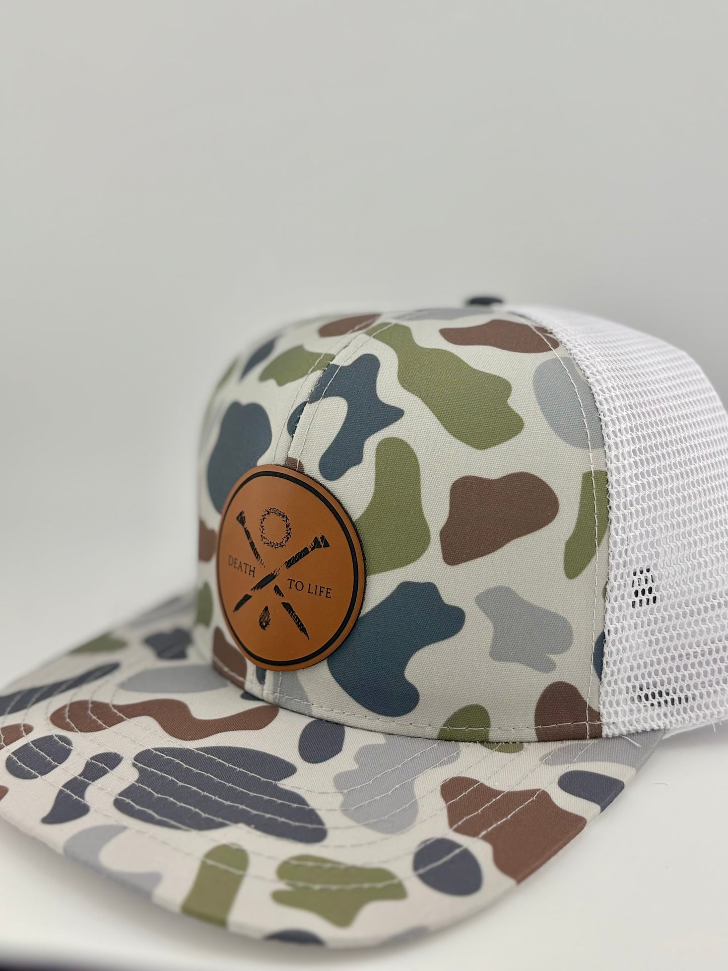 "Side view of a camo trucker hat with a structured front panel and a breathable white mesh back. The brown leather patch on the front showcases crossed arrows, a wreath, and the words 'Death to Life,' emphasizing a rugged yet stylish look."