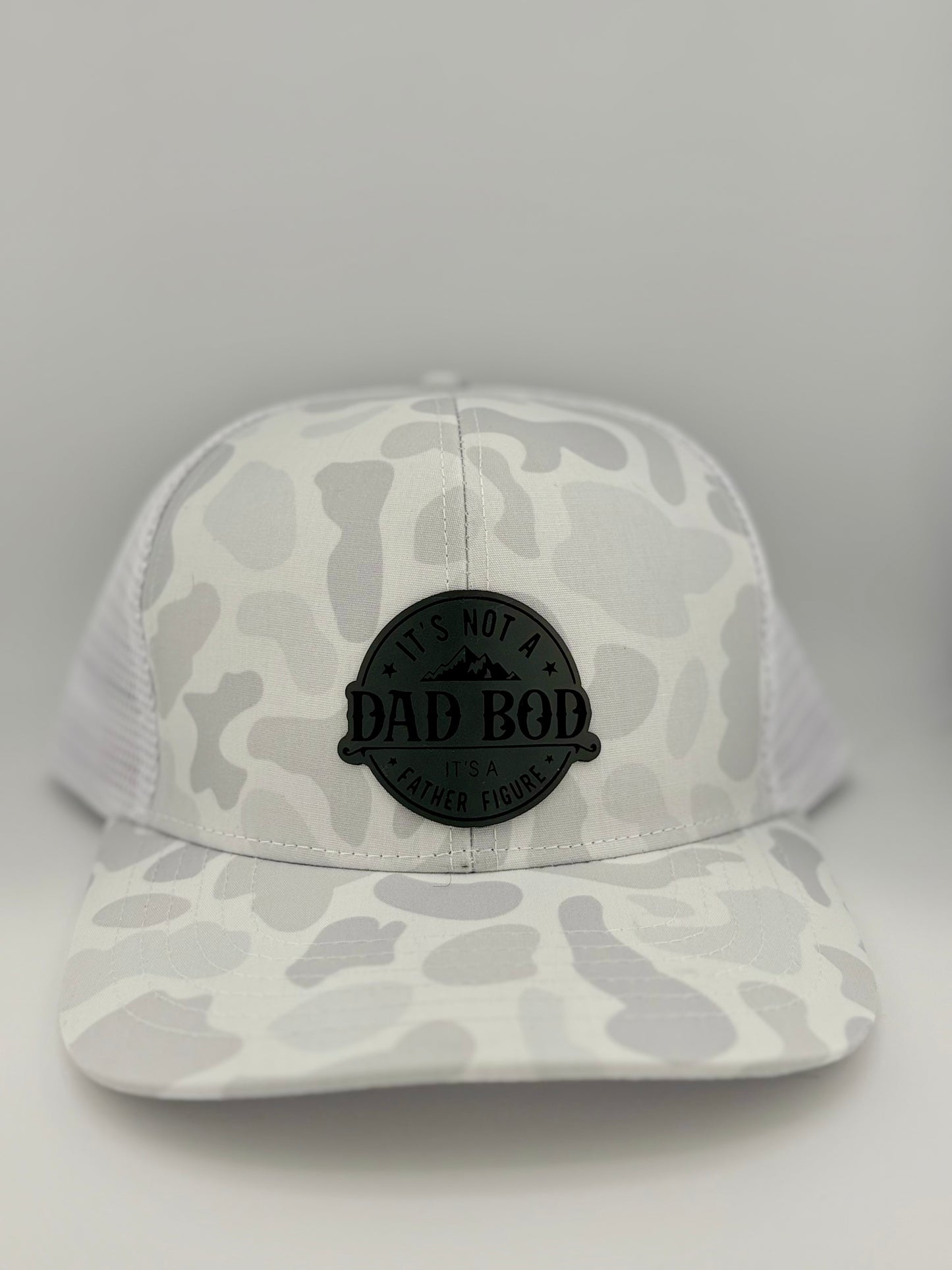 "White and light gray camo trucker hat with a structured front panel and a white mesh back. A black circular patch on the front displays the phrase 'IT'S NOT A DAD BOD, IT'S A FATHER FIGURE' with a mountain design. The monochrome color scheme adds a sleek and modern look."