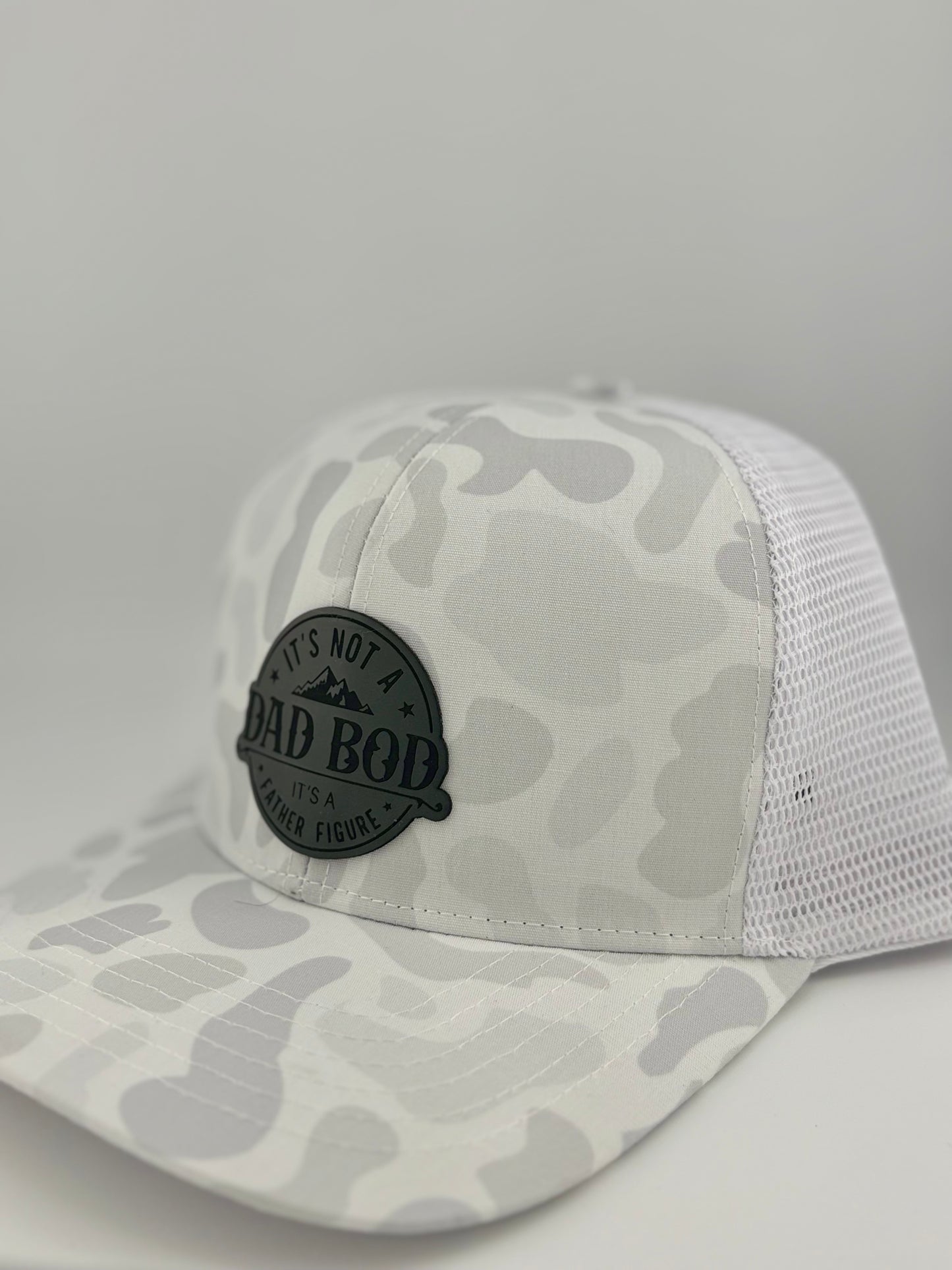 "Side view of a white camo trucker hat with a white mesh back and structured front panel. The black circular patch featuring 'IT'S NOT A DAD BOD, IT'S A FATHER FIGURE' and a mountain design adds a bold yet stylish statement for dads. The neutral colors make it a versatile accessory."