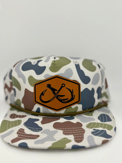 Camo-patterned snapback hat with a brown leather patch featuring an engraved double fishhook design inside a hexagon. The hat has an outdoor-inspired aesthetic with earthy tones of green, brown, and gray, complemented by a braided rope detail above the brim.