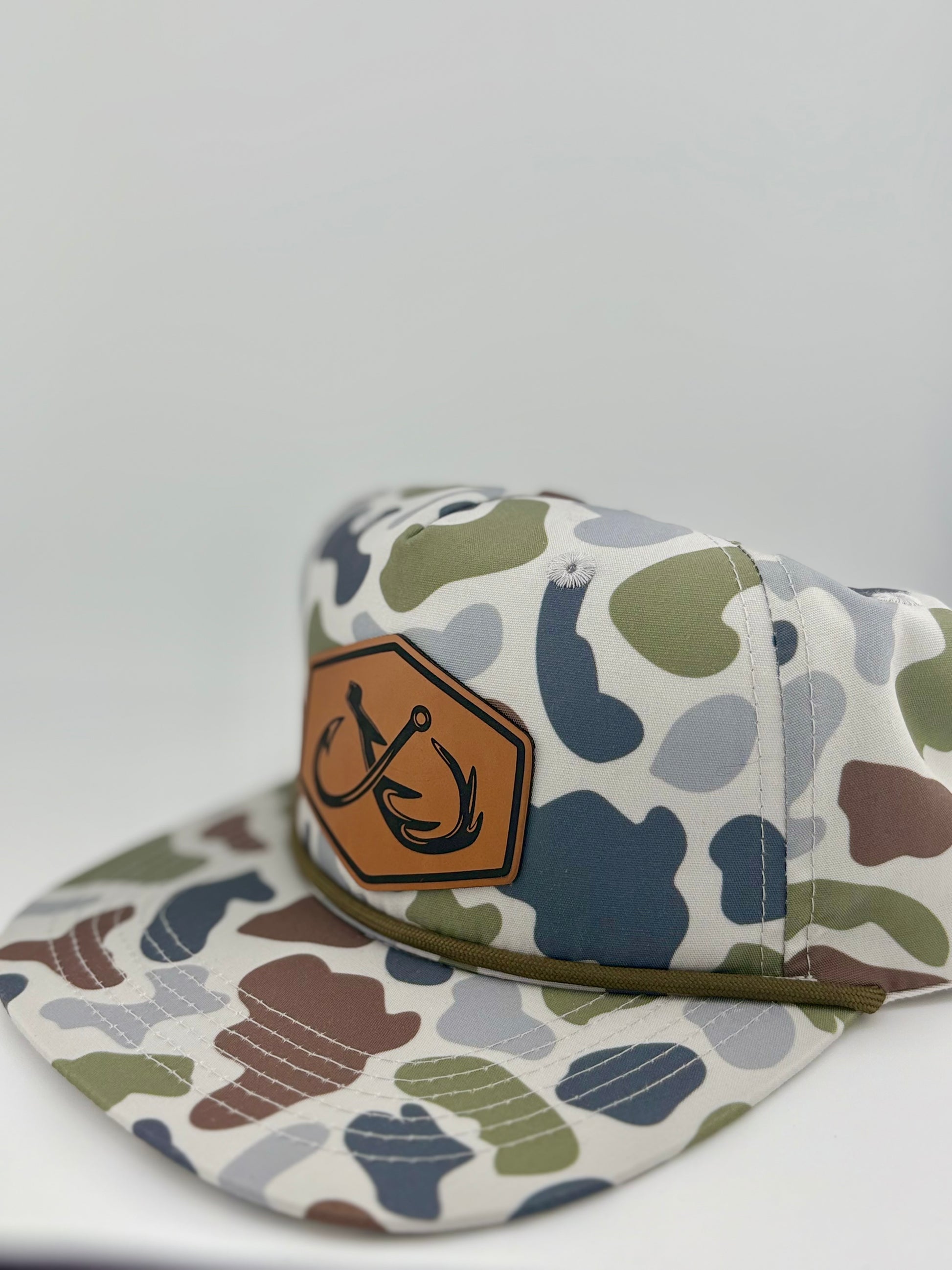 Camo-patterned snapback hat with a brown leather patch featuring an engraved double fishhook design inside a hexagon. The hat has an outdoor-inspired aesthetic with earthy tones of green, brown, and gray, complemented by a braided rope detail above the brim.